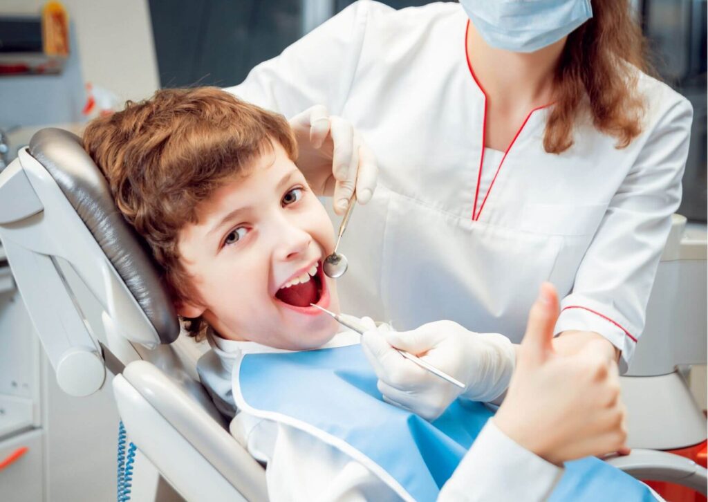 Young boy giving a thumbs-up during a dental visit while receiving care after losing his first baby tooth.